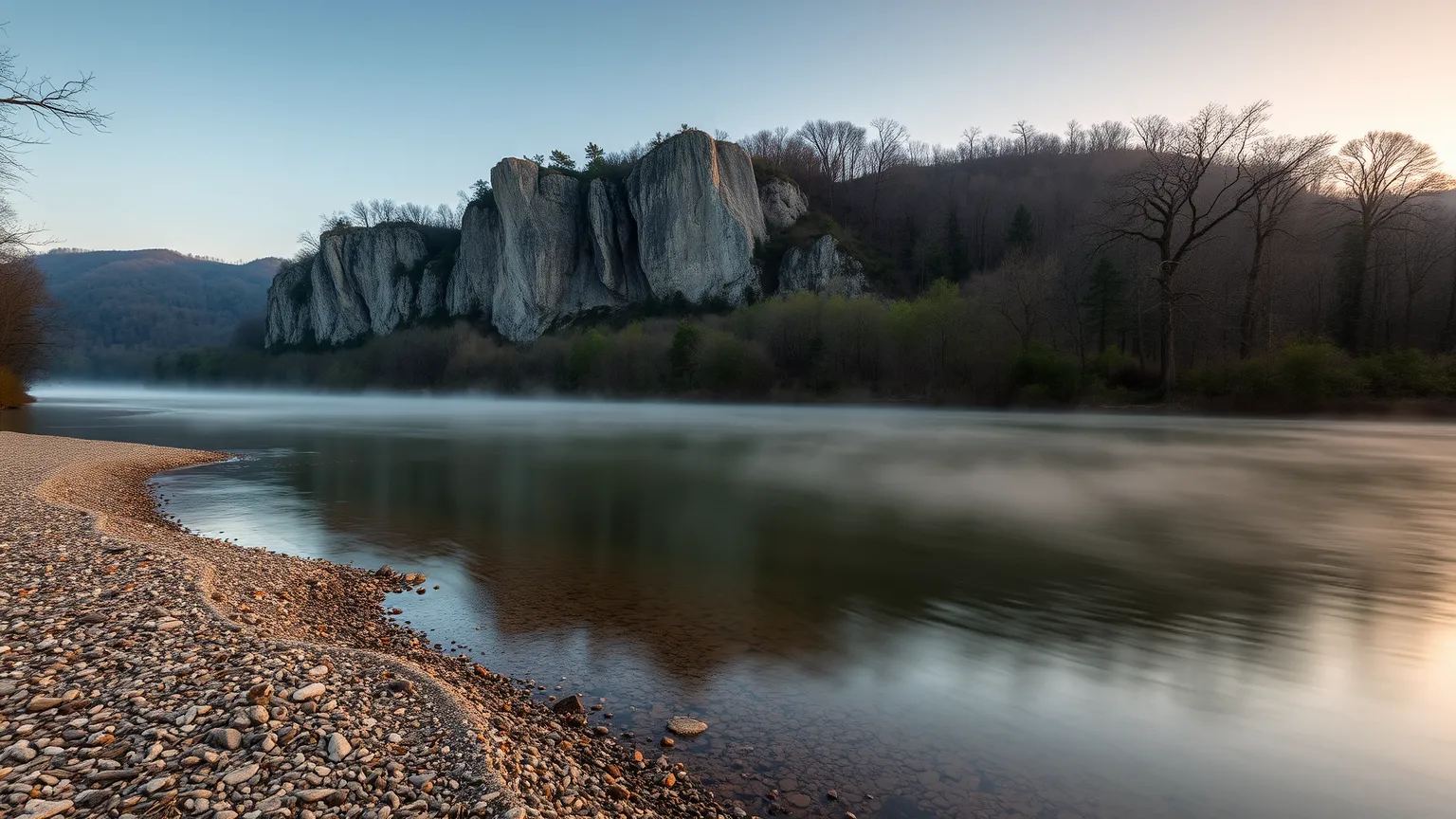 A calm, clear Ozarks river bend at dawn, with morning mist rising beneath a limestone bluff and forested hills.